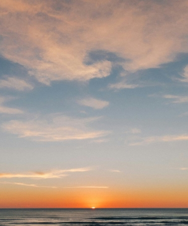 A serene sunset over the ocean with the sun just dipping below the horizon. The sky is painted with soft orange and pink hues, and wispy clouds are scattered across the upper part of the sky