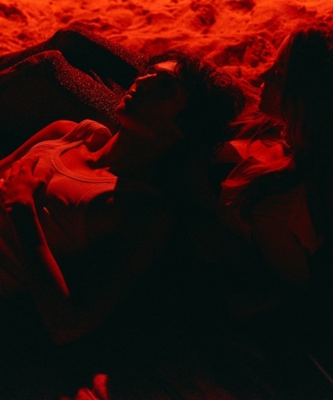 Two people lying on a beach at night under red lighting, creating a warm, intimate ambiance. One person, in a tank top, reclines with a relaxed posture, gazing up, while the other leans closely beside them. The surrounding sand and blankets are also bathed in the deep red glow, adding to the scene’s atmospheric and contemplative mood.