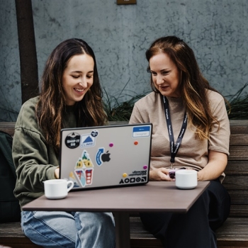 Two attendees collaborate outdoors at Design Leadership 2025, smiling over a laptop covered in stickers, with coffee cups resting on the table.