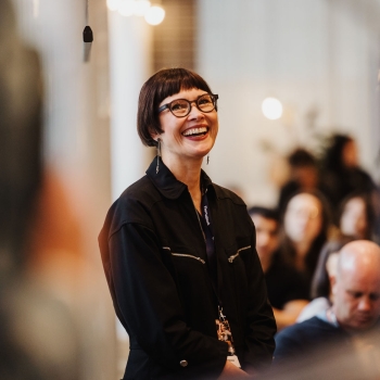 Smiling attendee in glasses and a black jumpsuit stands in a warmly lit room at The Outlook, with a softly focused audience in the background.