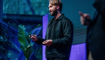 Oliver Ree giving a presentation at DO22, wearing a black jacket and using a microphone headset, with a leafy plant and large windows in the background