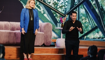 Trudi (left) and Mark (right) addressing the audience on stage at a Design Outlook event. Trudi is wearing a bright blue blazer and black outfit, while Mark, dressed in black, is actively signing as an Auslan interpreter. The stage features a modern setup with a purple sofa and decorative flowers, framed by large glass windows revealing greenery outside.