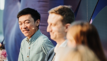 Xile Wang, wearing a checkered shirt and a headset microphone, smiling warmly during a panel discussion at a Design Outlook event. He is seated alongside other panelists, with the foreground slightly blurred, creating a focus on his engaging expression. The background features soft tones and abstract shapes, complementing the professional atmosphere.