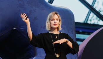 MC Monsalve presenting at a Design Outlook event, gesturing with one hand raised while holding a clicker in the other. She is wearing a black outfit with a statement gold necklace and a headset microphone, with a confident and poised expression. The backdrop features abstract blue shapes and modern architectural elements, enhancing the event’s contemporary ambiance.