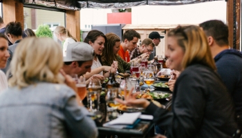 Large group of people gathered at a long outdoor table, enjoying food and drinks together in a lively, social dining setting.