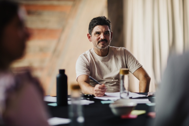 A man with short dark hair and a beard, wearing a light-coloured T-shirt, sitting at a table during the Design Leadership event, holding a pen and looking thoughtful