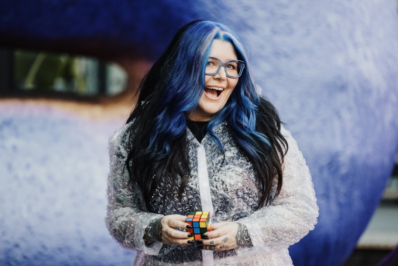 Chi Ryan, with vibrant blue and black hair and blue glasses, wearing a transparent bubble wrap coat, smiling enthusiastically while holding a Rubik’s Cube. She stands against a soft-focus background of abstract blue shapes, exuding a playful and dynamic vibe.