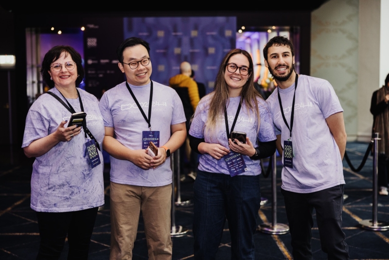 Four volunteers at the DO 2024 conference stand together, smiling and holding their phones. They are all wearing matching purple DO24 event t-shirts with lanyards and badges. The group appears cheerful and ready to assist attendees. Behind them, a backdrop with the conference branding is visible, along with a few attendees in the background.