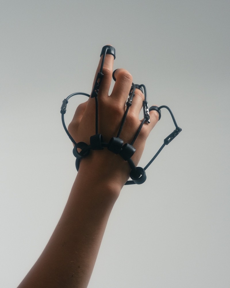 A close-up of a hand wearing mechanical attachments on each finger, connected by black cords, against a light, minimalistic background, evoking a modern, technology-inspired feel.