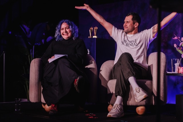 DO24 panelists seated on stage, one smiling with notes in hand, while the other raises his arms expressively, creating an engaging and dynamic moment.