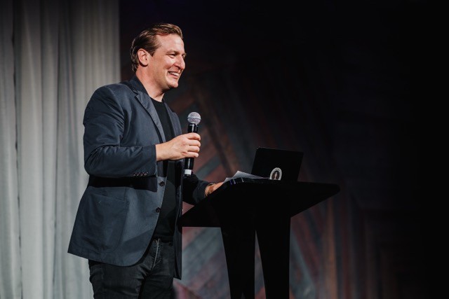 Mickey Rummery speaks confidently on stage at a DesignOutlook event, smiling and engaging the audience while holding a microphone.