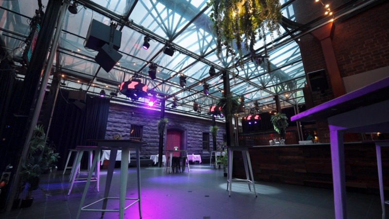 Royal Melbourne Hotel’s glass-roofed atrium set up for an event, featuring moody lighting, greenery, and cocktail tables under a vaulted ceiling.