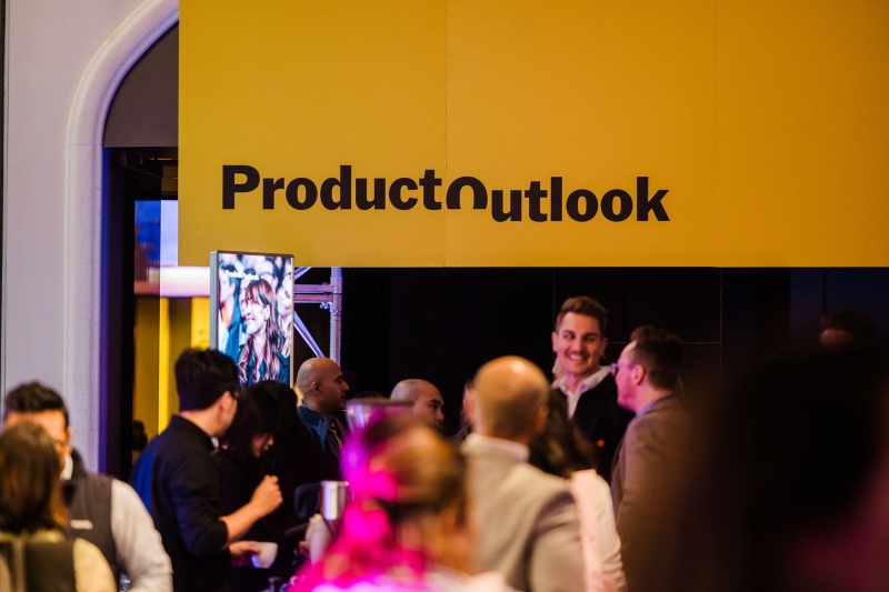 Crowd gathered beneath bright yellow Product Outlook signage at Product Outlook 2025 in Melbourne, with attendees chatting and a digital screen nearby.