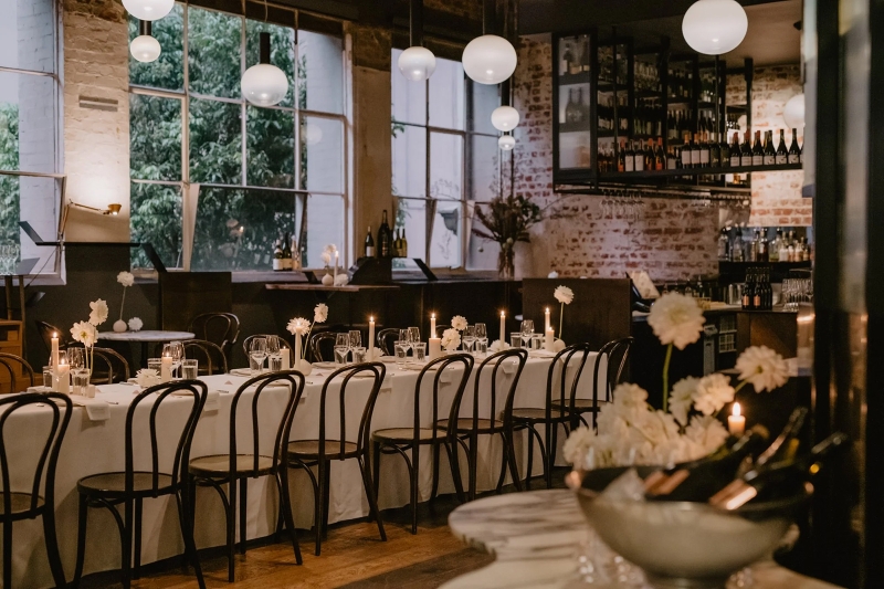 Elegant dinner setting at Cumulus Inc, with long white-clothed tables, candles, wine glasses, and minimalist floral arrangements in a warm, rustic interior.