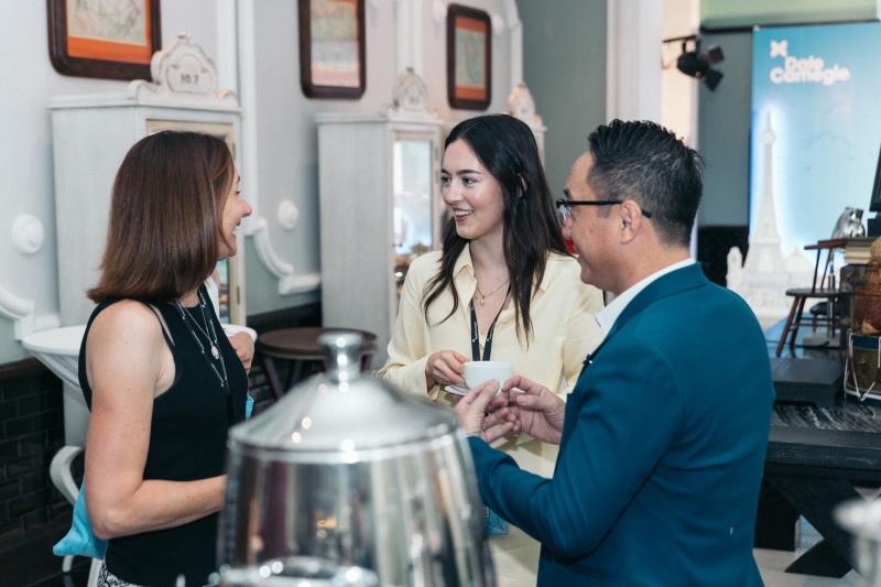 Three people stand together at a networking event, smiling and chatting while holding coffee cups, with a Dale Carnegie banner visible in the background.