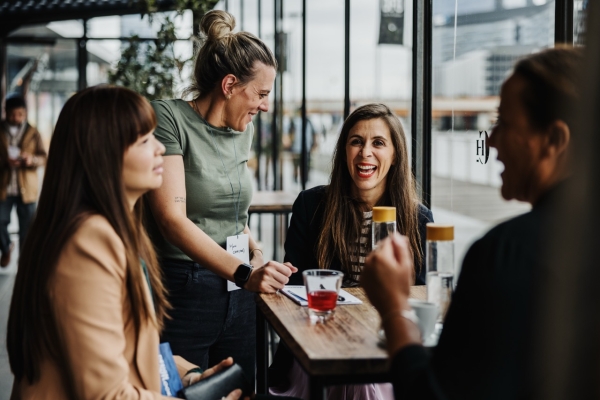 A lively moment in a café-like setting with four women chatting around a wooden table near large windows. One stands smiling in a green t-shirt, while another seated in a striped top and blazer laughs warmly. The setting feels modern and friendly, with an urban view outside.