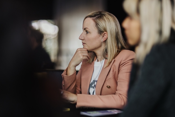 A woman with blonde hair, wearing a light peach blazer over a white top, sits thoughtfully with her hand resting on her chin. She appears focused, looking towards the right side of the frame. The background is softly blurred, emphasizing her contemplative expression during what seems to be a conference or meeting setting.