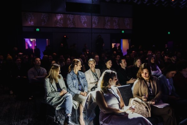 DO24 audience immersed in a session, with front-row attendees illuminated by stage lighting, engaged in conversation and note-taking in a dimly lit room.