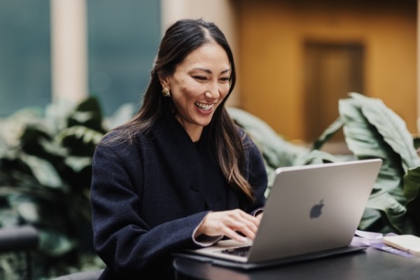 A DO24 attendee smiles while working on a laptop in an outdoor setting, surrounded by lush greenery, capturing a moment of focus and enjoyment.