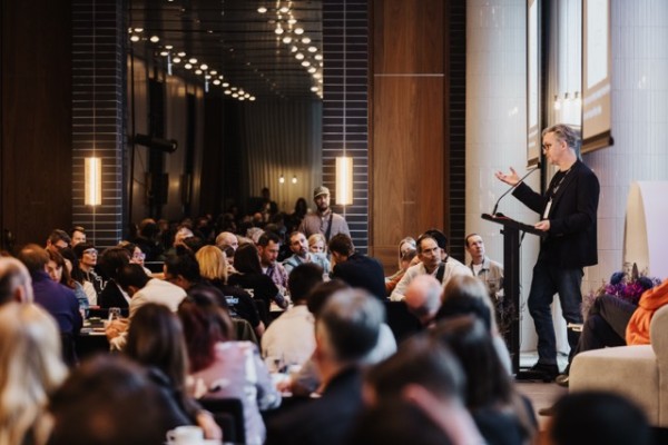 A speaker addresses a full room at a professional event. Attendees sit at round tables, listening closely in a modern, well-lit conference space.