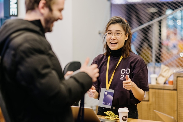 Evon Tan, wearing a DO22 volunteer shirt and lanyard, enthusiastically welcomes an attendee at the registration desk with a smile and thumbs up.
