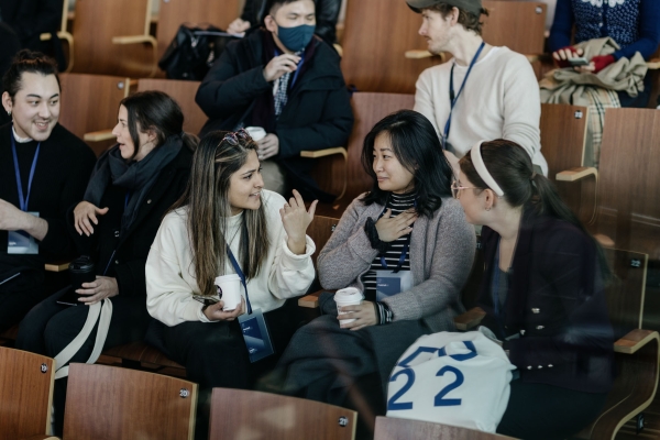Attendees at Design Outlook chat and sip coffee between sessions. They’re seated in wooden auditorium chairs, wearing conference lanyards and winter layers.
