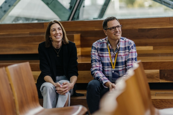 Presenters Alice King and John Lazarra sit in a timber-seated venue, smiling. Alice wears a black blazer, John a plaid shirt with a yellow lanyard.