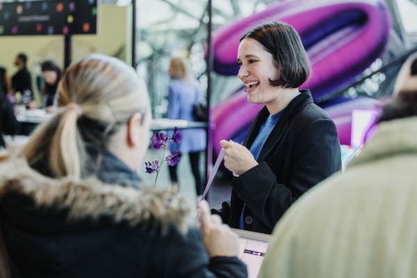 Smiling event staff member at Design Outlook 2023 hands a lanyard to an attendee at the check-in desk, with purple signage and orchids nearby.