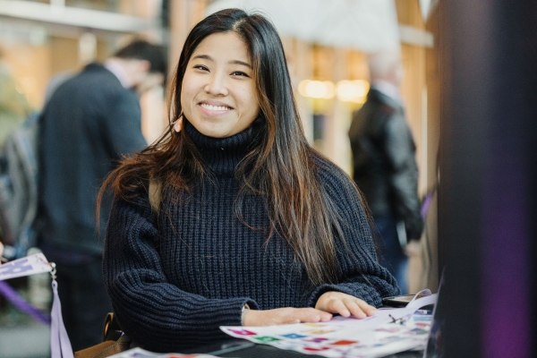 Smiling attendee at the check-in desk holding a printed program at Design Outlook, with people and venue lighting softly blurred in the background.