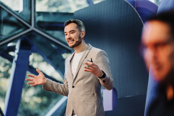 Presenter, Jonathan Lau, on stage at Design Outlook 2023, speaking with animated gestures while wearing a grey blazer and headset mic.