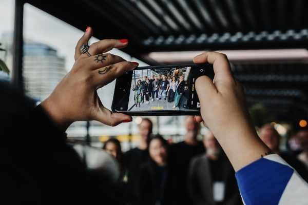Close-up of hands holding a phone, capturing a group photo of smiling event attendees outdoors.