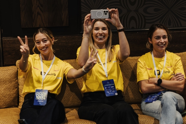 Miro team members at Design Outlook 2024 smiling on a couch, flashing peace signs and capturing moments while repping their bright yellow event shirts.
