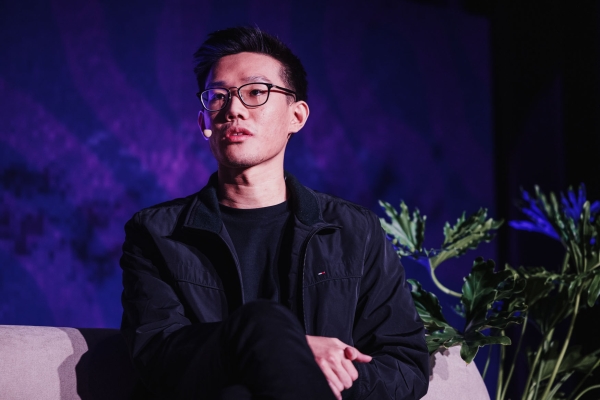 Felix Lee sits on stage during his Design Outlook 2024 presentation. Wearing glasses and a black jacket, he speaks thoughtfully into a headset mic against a vibrant purple backdrop, with leafy plants softly lit beside him.