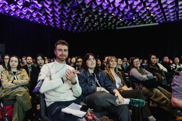 A full room of attendees at Design Outlook 2024, listening attentively, smiling, and clapping under the event’s signature purple lighting.
