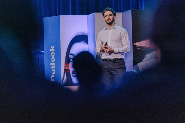 Male speaker framed by audience silhouettes (file-Kpwb53kzw1UsPpWBg2HLVj) Male speaker stands mid-presentation on stage at The Outlook, framed by silhouettes of attendees and panels with striking visual motifs.