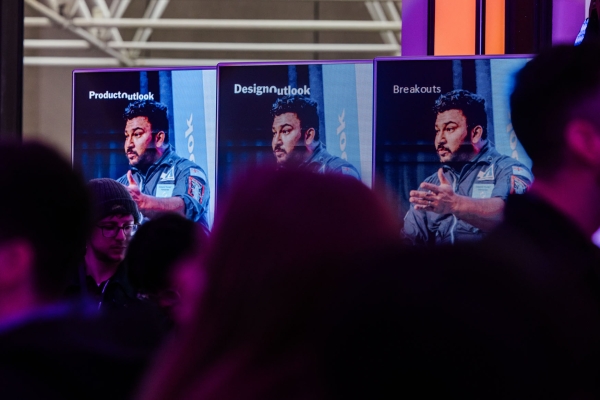 Multiple screens at The Outlook display speaker Dwayne Fernandes in a flight suit, with session titles ‘ProductOutlook’, ‘DesignOutlook’, and ‘Breakouts’.