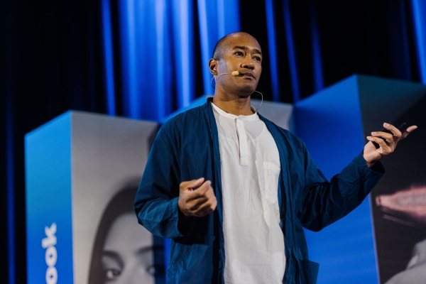Corey Lee presenting on stage at Design Outlook 2025, wearing a headset mic and gesturing mid-talk against the event’s bold blue backdrop.