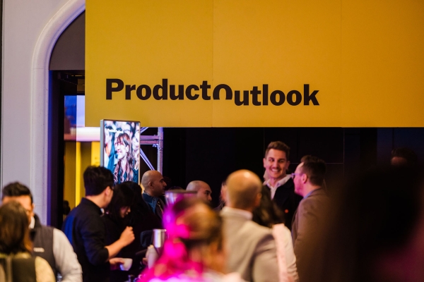 Crowd gathered beneath bright yellow Product Outlook signage at Product Outlook 2025 in Melbourne, with attendees chatting and a digital screen nearby.