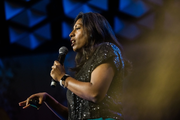 Presenter Sathya Bala presenting on stage at Product Outlook 2025 in Melbourne, holding a microphone and clicker, framed by dramatic lighting and geometric ceiling panels.