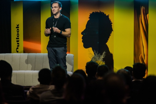 Presenter Matt Perrott speaking on stage at Product Outlook 2025 in Melbourne, standing in front of The Outlook’s signature yellow panels and a silhouette backdrop, addressing an engaged seated audience.