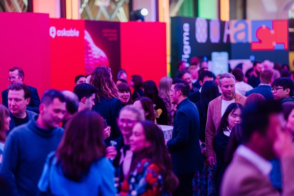 Colorful crowd scene at TO25, with attendees networking and chatting in front of bright sponsor displays under vibrant purple and pink lighting.