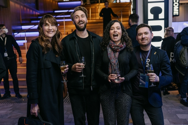 Four people stand together at an event, smiling and holding drinks, with a warmly lit staircase and “WELCOME” signage in the background.