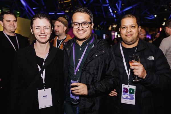 Three attendees wearing event lanyards smile at the camera while holding drinks during a lively evening gathering under purple lighting.