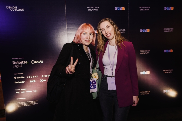 Two attendees pose and smile at the Design Outlook event backdrop, one flashing a peace sign, both wearing event lanyards and stylish outfits.