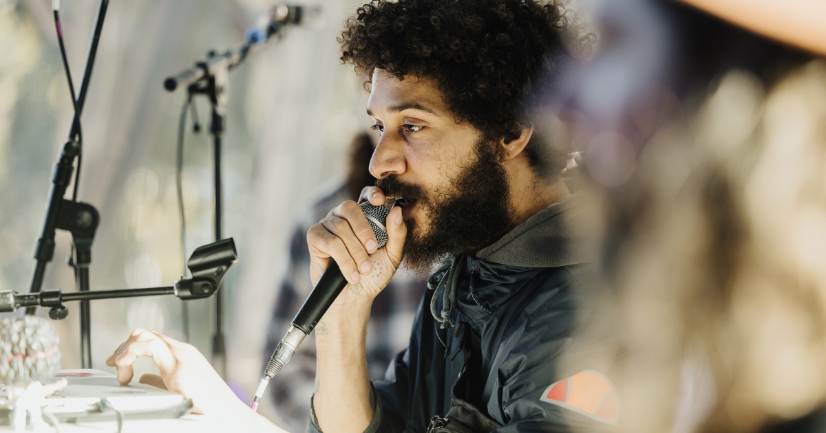 A performer at a Design Outlook event, holding a microphone close to their mouth while interacting with equipment on a table. They have curly hair, a beard, and are wearing a dark jacket. Microphone stands and blurred details in the background create a dynamic and creative setting, capturing the energy of the moment.