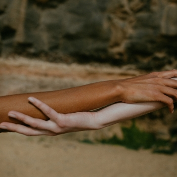 Two people holding each other’s forearms gently, with one person having a darker skin tone and the other a lighter skin tone, set against a natural background