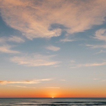 A serene sunset over the ocean with the sun just dipping below the horizon. The sky is painted with soft orange and pink hues, and wispy clouds are scattered across the upper part of the sky