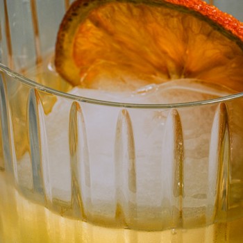 A close-up of a refreshing cocktail in a textured glass, featuring a large ice cube and a dried orange slice garnish. The golden tones of the drink and the intricate reflections on the glass create a warm and inviting aesthetic.
