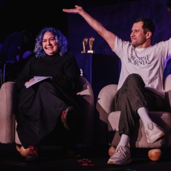 DO24 panelists seated on stage, one smiling with notes in hand, while the other raises his arms expressively, creating an engaging and dynamic moment.