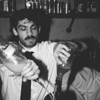 A bartender in black-and-white, focusing intently as he pours drinks from two bottles into a shaker during a busy service behind a bar.